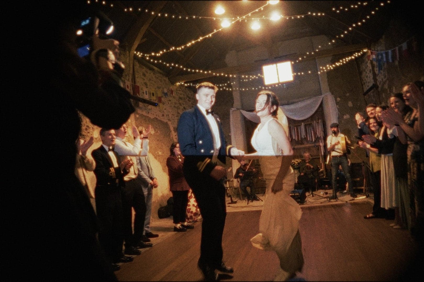 A couple dancing between clapping guests in a barn strung with fairy lights, seen past a camera in the foreground